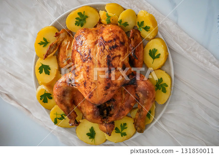 Close-up of a whole roasted chicken surrounded by round potatoes on a festive holiday table. Cozy Christmas, Thanksgiving, or New Year family celebration with colorful and appetizing food presentation 131850011
