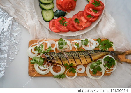 Close-up of a festive holiday presentation featuring herring garnished with onion rings on a wooden board, accompanied by a plate of sliced tomatoes and cucumbers. Cozy Christmas, New Year, or Close-up of a festive holiday presentation featuring herring garnished with onion rings on a wooden board, accompanied by a plate of sliced tomatoes and cucumbers. Cozy Christmas, New Year, or 131850014