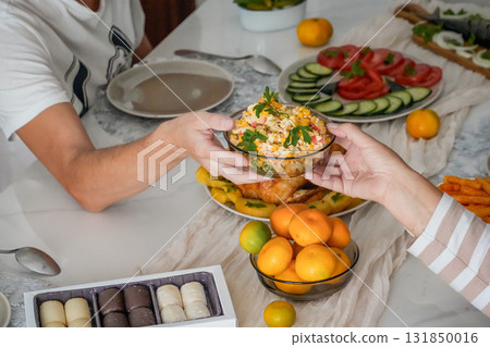 Close-up of hands holding a bowl of Olivier salad above a festive holiday table with roasted chicken, mandarins, and sweets. Cozy Christmas and New Year celebration with traditional dishes and warm 131850016