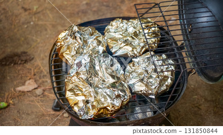 Grilled food wrapped in foil is cooking on a charcoal barbecue grill, surrounded by natural outdoor setting, showcasing summer cooking experience 131850844