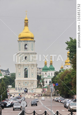 View of St. Sophia's Cathedral bell tower with golden domes in Kyiv, Ukraine, captured from street level with cars and pedestrians in the foreground on a cloudy day. 131851732