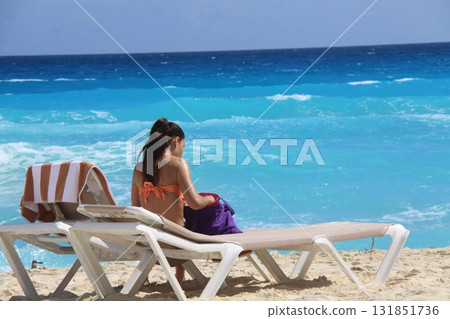 Woman in an orange bikini sitting on a beach lounger facing the turquoise sea 131851736