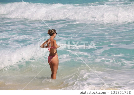 Woman in a pink bikini stands in shallow turquoise surf, adjusting her top while facing the ocean waves, enjoying a sunny day at a tropical beach Woman in a pink bikini stands in shallow turquoise surf, adjusting her top while facing the ocean waves, enjoying a sunny day at a tropical beach 131851738