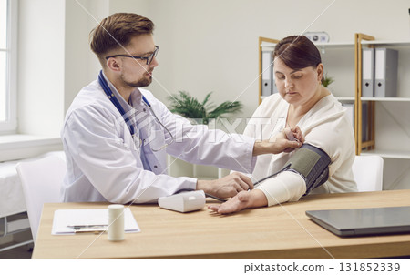 Male doctor using sphygmomanometer to measure blood pressure of young overweight woman Male doctor using sphygmomanometer to measure blood pressure of young overweight woman 131852339