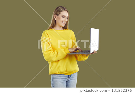 Smiling blondy woman in casual yellow sweatshirt and jeans working on laptop on khaki background. Smiling blondy woman in casual yellow sweatshirt and jeans working on laptop on khaki background. 131852376