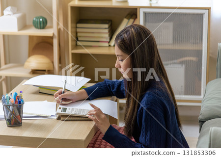 Teen student girl does homework, writes notes for study using calculator, prepares for exam at school desk, young thailand woman shows serious learning focus with focused expression. Teen student girl does homework, writes notes for study using calculator, prepares for exam at school desk, young thailand woman shows serious learning focus with focused expression. 131853306