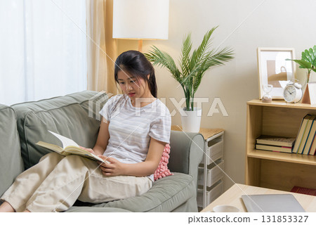Young woman reading book, teen person relaxing on sofa, finding rest in cozy room, home environment offers comfort, pleasant bright interior design features green plant, wooden shelf, hourglass. Young woman reading book, teen person relaxing on sofa, finding rest in cozy room, home environment offers comfort, pleasant bright interior design features green plant, wooden shelf, hourglass. 131853327