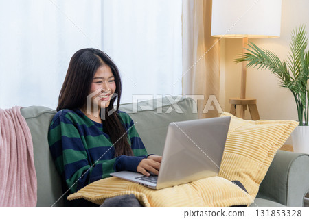 Happy teen woman uses laptop at home for work, shopping or customer connection, smiling and relaxing on plush cozy sofa, wearing warm striped shirt, enjoying computer activity. Happy teen woman uses laptop at home for work, shopping or customer connection, smiling and relaxing on plush cozy sofa, wearing warm striped shirt, enjoying computer activity. 131853328