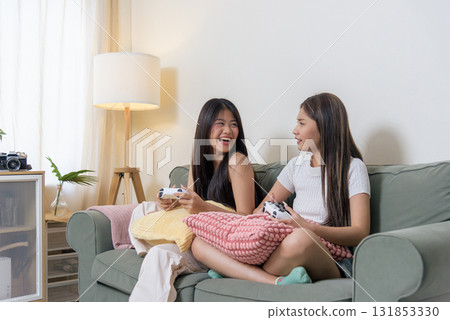 Two young women friends play exciting console video game home, teenagers enjoy playing popular videogame together with joy and laughter, creating fun memorable moments in cozy living room setting. 131853330