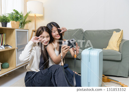 Teen friends packing luggage, happy girls prepare bags for travel vacation, showing excitement for trip preparation, holding camera and making peace signs, looking forward to fun adventure. 131853332