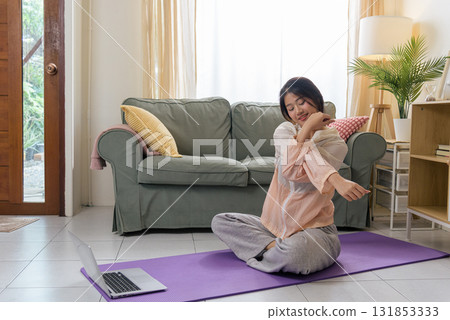 Healthy lifestyle woman exercises yoga for physical health, teen workout at home with sport, young Asian woman stretches arm on purple mat watching online fitness on laptop in bright room. 131853333
