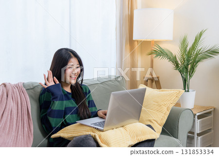 Happy teen woman uses laptop at home, enjoying work, customer shop tasks, daily use, relaxing on living room sofa, waving to device, casual outfit, bright daylight illuminates cozy indoor space. 131853334