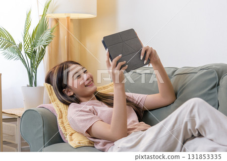 Teen person using tablet, woman relaxing reading digital content, enjoying entertainment, modern communication, happy teen girl smiling at device screen, leisure activity at home sofa. 131853335