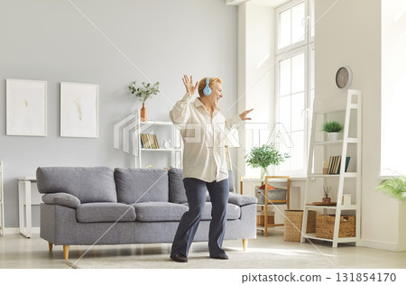 Cheerful mature woman with blonde hair wearing headphones dancing in bright modern living room. Cheerful mature woman with blonde hair wearing headphones dancing in bright modern living room. 131854170