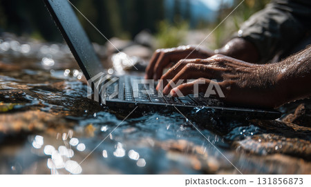 person is typing quickly on waterproof laptop near river, surrounded by nature. scene captures essence of working outdoors, showcasing beauty of technology and nature together 131856873