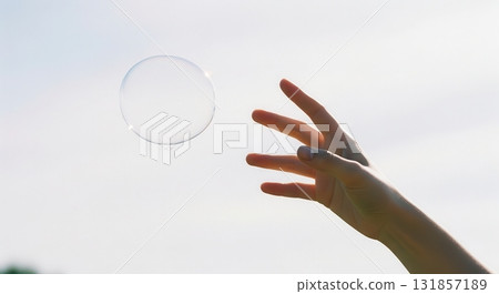 Hand reaching out to catch a delicate soap bubble floating in the bright sky, capturing a moment of playful joy 131857189
