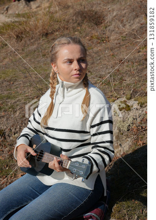 Beautiful blonde girl playing guitar ukulele and singing relaxing enjoying outdoors on sunny autumn day. Hippie woman in boho style training playing musical instrument. Fall leaves 131859182