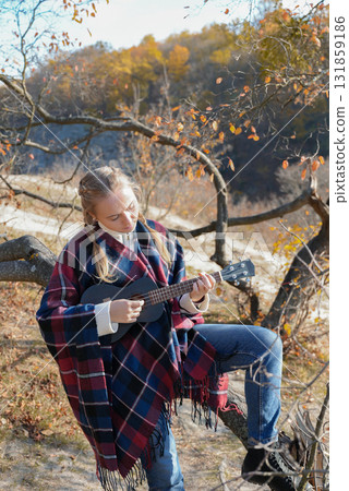 Blonde woman playing guitar ukulele outdoors in autumn forest. Concept of sound therapy, mental health and wellness rituals. Calmness tranquility audio-sensory practices. Aura farming 131859186