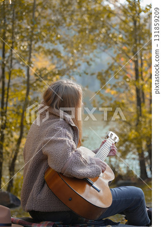 Blonde woman playing string guitar outdoors in autumn forest. Concept of sound therapy, mental health and wellness rituals. Calmness tranquility audio-sensory practices. Aura farming energy 131859209