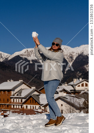 Snowball Fight Mountains Winter Fun: Woman playfully throws snowball near snowy village, sunny day. 131861356