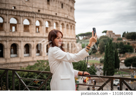 Woman in white blazer and red dress taking selfie with smartphone near Colosseum in Rome 131861608