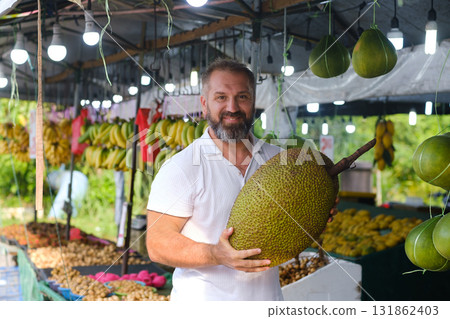 Man holding large jackfruit at a vibrant outdoor fruit market in tropical region during sunny daytime 131862403