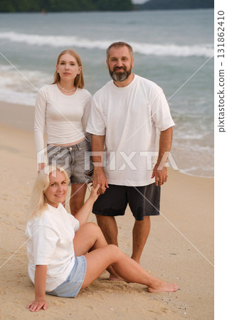 Family enjoys a sunny day at the beach, smiling while standing together by the shoreline with gentle waves in the background 131862410