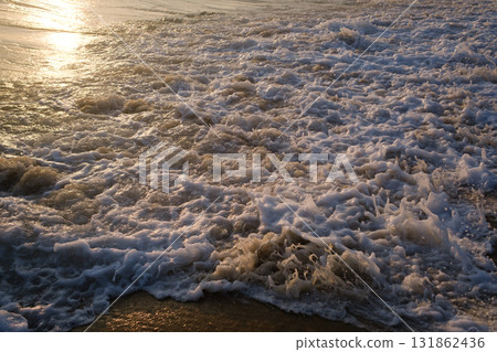 Sunset view of waves crashing on the beach with silhouetted figures enjoying water activities in the distance near an island Sunset view of waves crashing on the beach with silhouetted figures enjoying water activities in the distance near an island 131862436