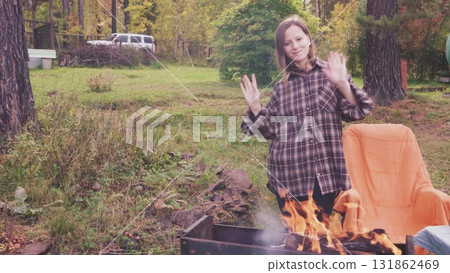A woman relishes a lovely autumn day outdoors while having a picnic next to a crackling fire. Surrounded by vibrant fall foliage, she captures the essence of the season with a cozy atmosphere. 131862469