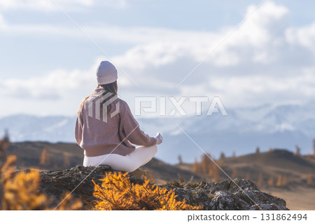 A woman sits gracefully on a rock, meditating in a serene mountain setting filled with autumn hues. The peaceful atmosphere enhances her yoga practice amidst nature. A woman sits gracefully on a rock, meditating in a serene mountain setting filled with autumn hues. The peaceful atmosphere enhances her yoga practice amidst nature. 131862494