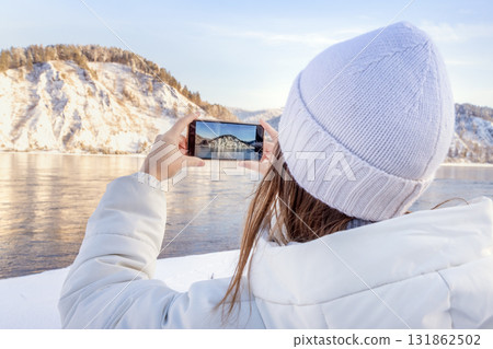 Capturing the breathtaking beauty of winter Scenic photography in a stunning snowy landscape, A woman photographing a stunning winter landscape near the calm waters of the Yenisei River near 131862502