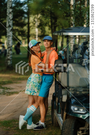 Couple enjoys a joyful moment near a golf cart in a lush park setting on a sunny day while dressed in vibrant golf attire 131862599