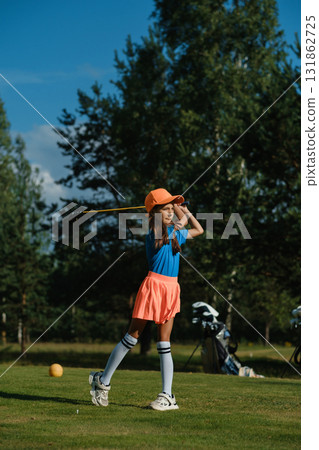 Young girl practicing golf swing on a sunny day at a green course surrounded by trees with sports equipment nearby Young girl practicing golf swing on a sunny day at a green course surrounded by trees with sports equipment nearby 131862725