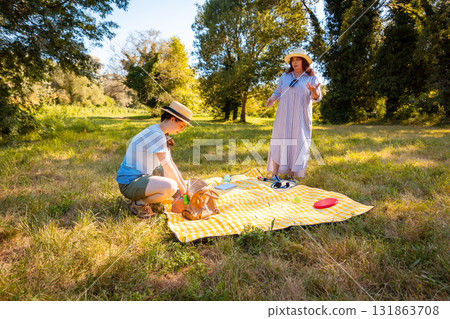 Middle-aged mother and her young Caucasian daughter relaxing on a blanket in a summer park enjoying healthy snacks and family recreation outdoors. Wide shot 131863708