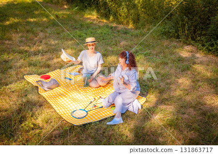 Two Caucasian women, mom and daughter, sitting on a yellow blanket in a summer park. Mental care, vacation, and connection concept. Two Caucasian women, mom and daughter, sitting on a yellow blanket in a summer park. Mental care, vacation, and connection concept. 131863717