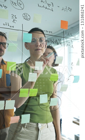 Professional businesswoman wearing glasses writing on glass board, marking strategic plan with sticky notes while colleagues brainstorming in background 131868241
