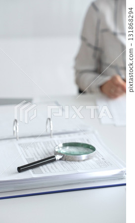 Magnifying glass on financial documents is in focus while an accountant using a calculator and taking notes, emphasizing accounting, finance, close up vertical view. Audit and taxes Magnifying glass on financial documents is in focus while an accountant using a calculator and taking notes, emphasizing accounting, finance, close up vertical view. Audit and taxes 131868294