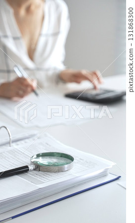 Magnifying glass on financial documents, with an accountant using a calculator and taking notes, emphasizing accounting, finance, and tax preparation, close up vertical view. Audit and taxes 131868300