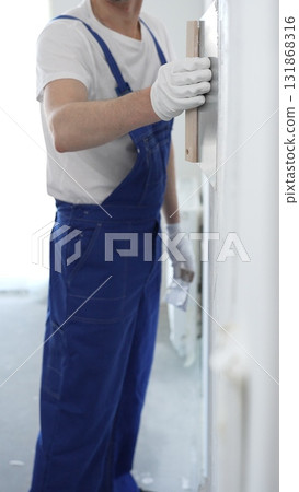 Man construction worker wearing protective gloves and blue construction coveralls, spreading plaster smoothly across wall using professional drywall taping knife Man construction worker wearing protective gloves and blue construction coveralls, spreading plaster smoothly across wall using professional drywall taping knife 131868316