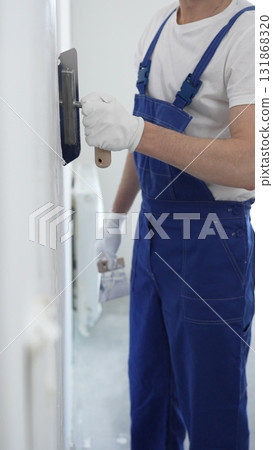 Professional construction worker wearing blue work overall is using a finishing trowel to carefully apply plaster on a wall, demonstrating expertise in home renovation Professional construction worker wearing blue work overall is using a finishing trowel to carefully apply plaster on a wall, demonstrating expertise in home renovation 131868320