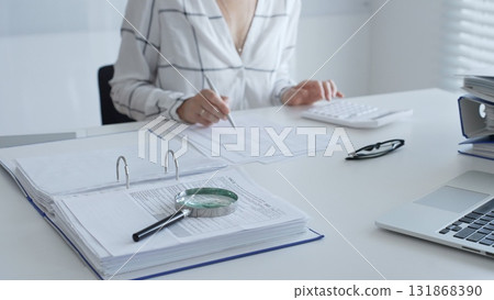 Female accountant is working with financial documents, using calculator and taking notes, with a magnifying glass and a folder on a white table in a bright office. Audit and taxes in business 131868390