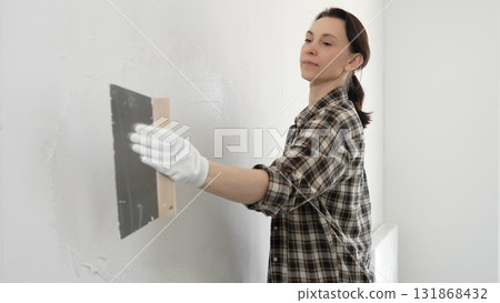 Woman construction professional wearing checkered shirt is smoothing white wall surface, applying putty with professional spatula technique, showcasing skilled interior renovation work 131868432