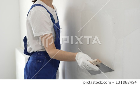 Unknown construction worker woman in blue overalls smoothing wall surface, applying plaster with putty knife, wearing protective white gloves during home renovation work Unknown construction worker woman in blue overalls smoothing wall surface, applying plaster with putty knife, wearing protective white gloves during home renovation work 131868454