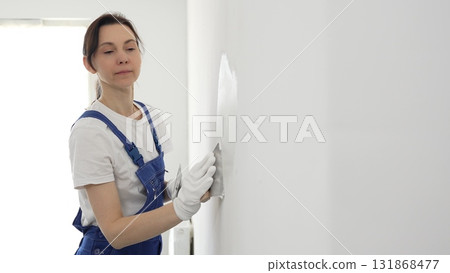 Construction worker woman spreading finishing putty on white wall, wearing protective gloves while smoothing surface with putty knife for precise home repair 131868477