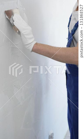 Close-up of man construction worker wearing gloves and blue overalls is using a putty knife to apply plaster on a white wall, performing home renovation and improvement 131868527