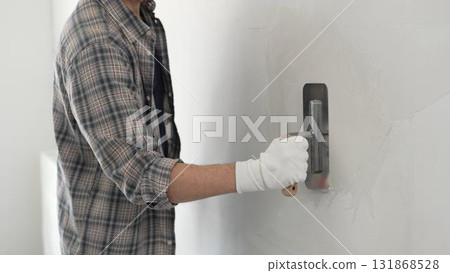 Unknown male construction worker wearing gloves and plaid shirt is using finishing trowel to plaster a white wall, applying a layer of plaster to create a smooth surface during renovation project 131868528