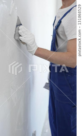 Close-up of man construction worker wearing gloves and blue overalls is using a putty knife to apply plaster on a white wall, performing home renovation and improvement Close-up of man construction worker wearing gloves and blue overalls is using a putty knife to apply plaster on a white wall, performing home renovation and improvement 131868529