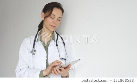 Doctor woman wearing white medical coat over green blouse, stethoscope, is reviewing patient records on digital tablet, standing in medical workspace with natural lighting. Medicine concept Doctor woman wearing white medical coat over green blouse, stethoscope, is reviewing patient records on digital tablet, standing in medical workspace with natural lighting. Medicine concept 131868538