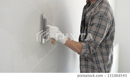 Unknown male construction worker wearing gloves and plaid shirt is using finishing trowel to plaster a white wall, applying a layer of plaster to create a smooth surface during renovation project Unknown male construction worker wearing gloves and plaid shirt is using finishing trowel to plaster a white wall, applying a layer of plaster to create a smooth surface during renovation project 131868543