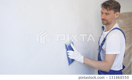 Man construction worker wearing protective white gloves and blue construction coveralls, is sanding white wall with blue hand block, preparing smooth surface during interior home renovation project 131868550